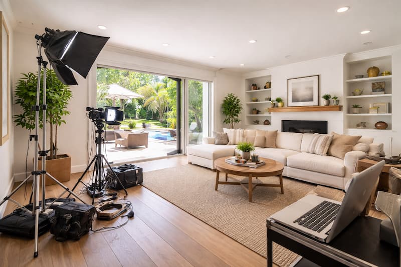Behind the scenes filming in a bright, modern living room with natural light, crew equipment, and a camera setup aimed toward the seating area